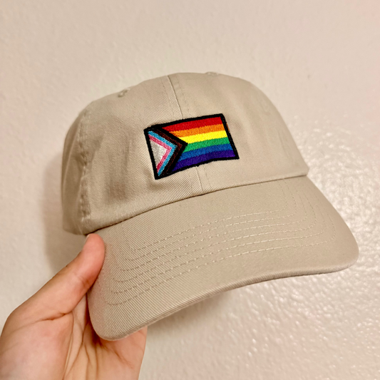 A hand holding a beige baseball cap with an embroidered Progress Pride flag patch on the front, set against a plain beige wall.